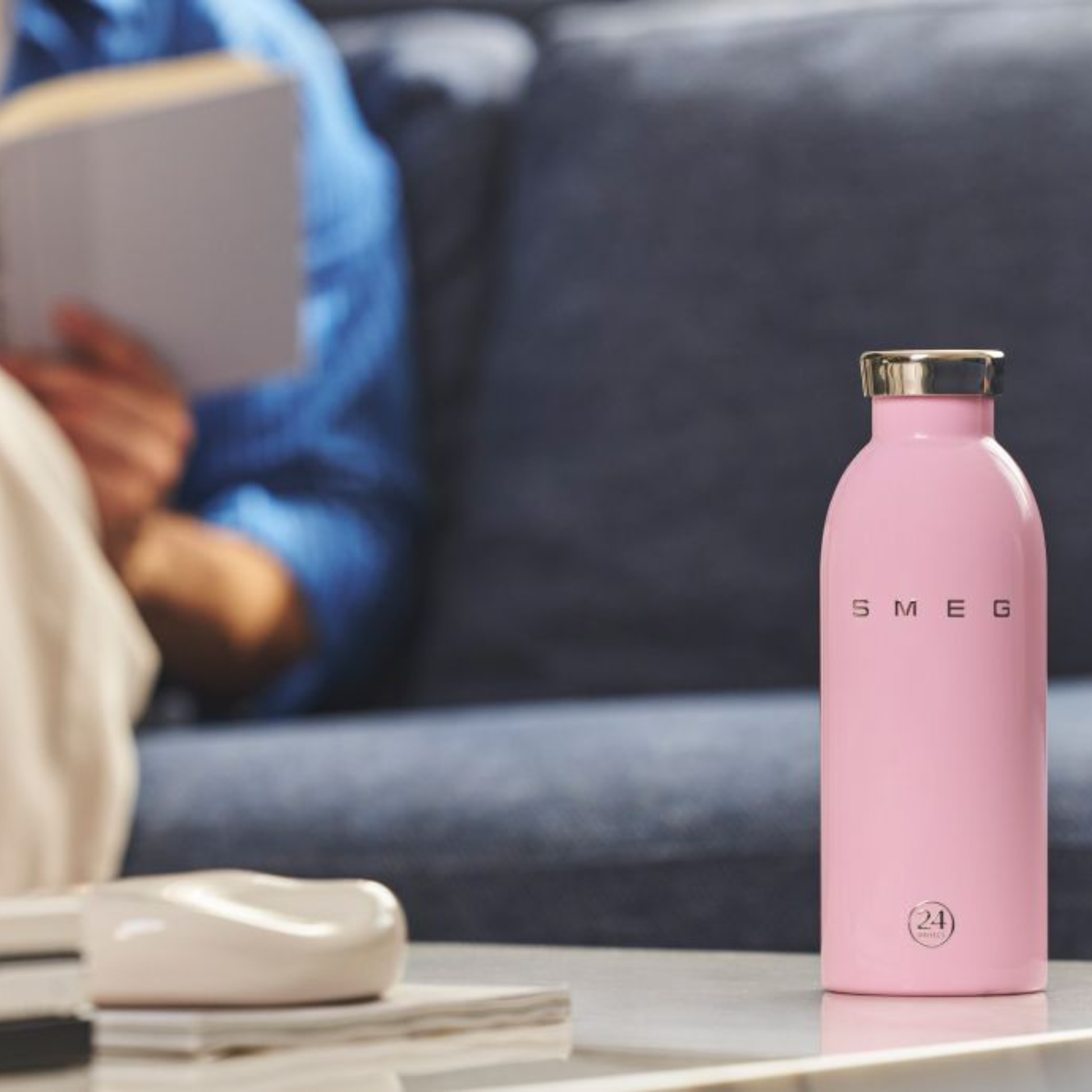 Pink water bottle with gold cap on a table, blurred person in blue shirt holding a book in the background